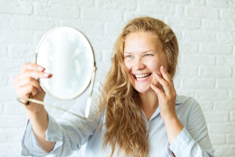 A blonde woman smiles while touching her face and looking in a small mirror after seeing a skin specialist in Minneapolis